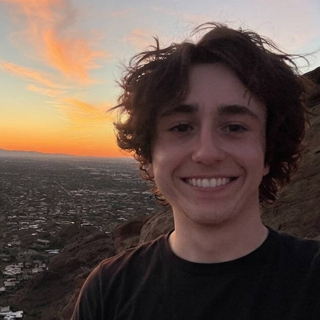 Smiling young man with curly brown hair in the foreground with a sunset horizon in the background